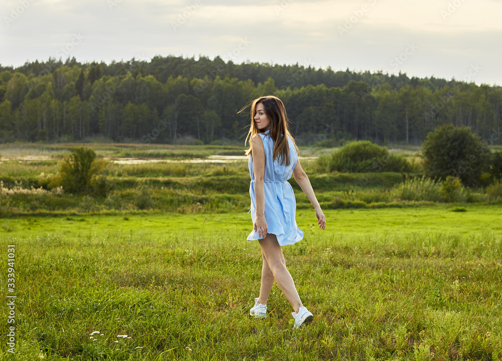 Young beautiful woman rushing into the wild nature: meadow, lake and forest, female in blue dress looking back and smiling, copy space, lifestyle, connection with nature concept