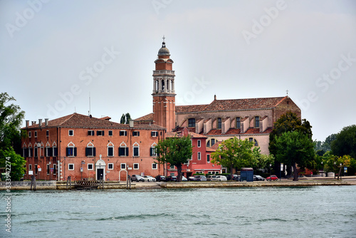 Venice, Italy - A brown building with a tiled roof of the church and monastery of San Nicolo del Lido, green trees on the shore near the water, gray sky in the summer in the daytime.