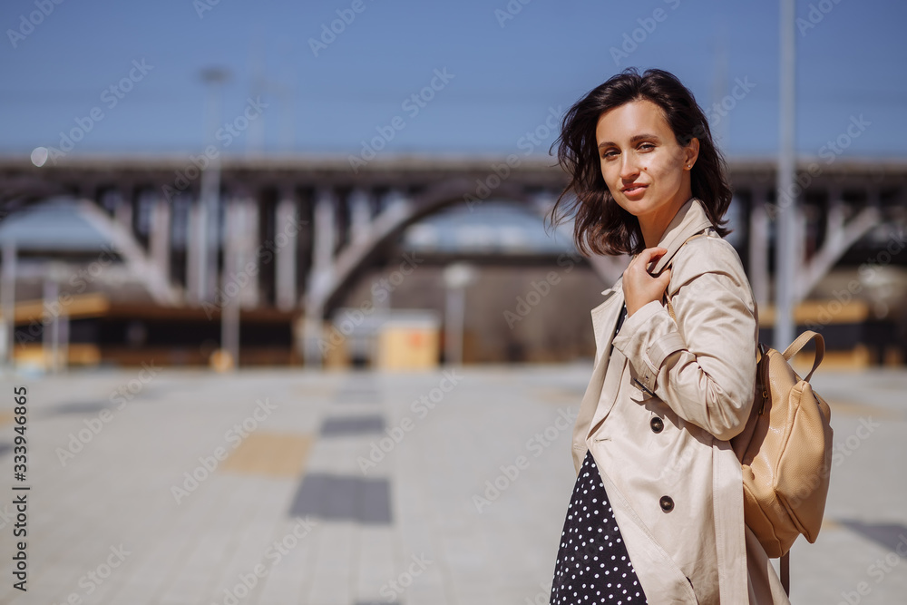 Fototapeta premium A beautiful girl model stands on the background of a bridge in the city in a long light cloak and a small backpack. Dress in peas. Urabinistic style.