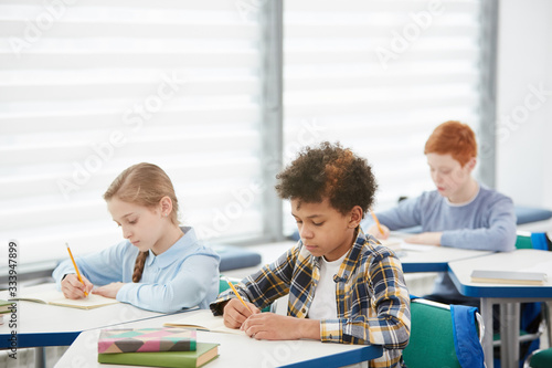Multi-ethnic group of kids sitting at desks in school classroom and writing or taking test, copy space © Seventyfour