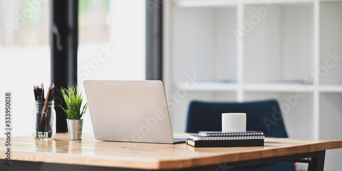 Photo of notebook, diary, coffee cup, pencil in vase, potted plant and computer laptop putting together on wooden working desk over modern living room windows and bookshelf as background. © Prathankarnpap
