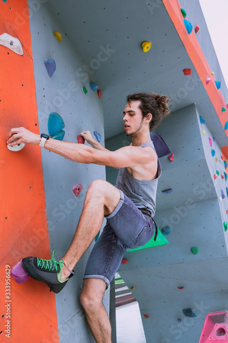 Urban concept of man with curly long hair, tied in a ponytail, is training at the city artificial red and blue climbing wall using talcum powder, wearing a bag for climbing without insurance equipment