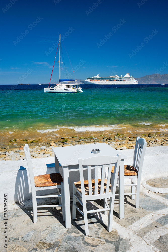 Tourist greek scene - restaurant cafe table on quay promenade with catamaran yacht and cruise liner and Aegean sea in background on beautiful summer day. Chora, Mykonos island, Greece
