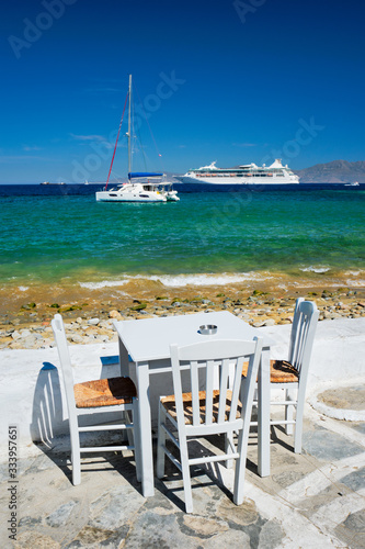 Tourist greek scene - restaurant cafe table on quay promenade with catamaran yacht and cruise liner and Aegean sea in background on beautiful summer day. Chora, Mykonos island, Greece © Dmitry Rukhlenko