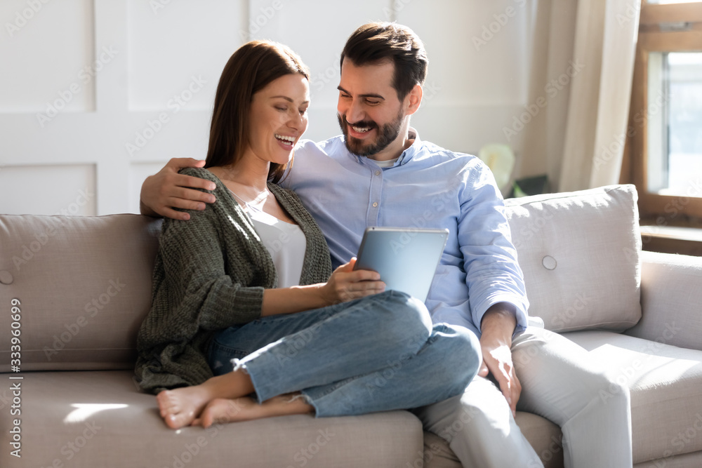 Excited young married couple resting on sofa, watching comedian funny movie on computer tablet, enjoying free weekend leisure time together. Happy family spouses cuddling, shopping online at home.