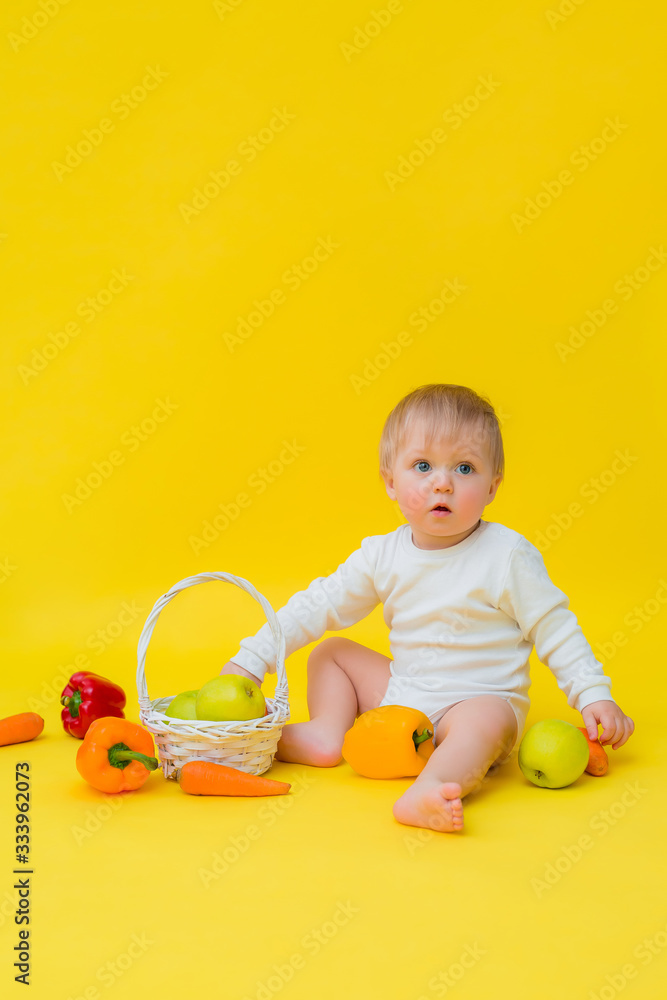 baby in a white bodysuit with vegetables, sitting on a yellow background. healthy food for the child, space for text baby in a white bodysuit with vegetables, sitting on a yellow background. healthy food for the child, space for text