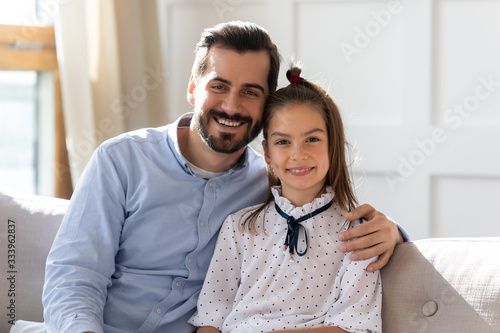 Head shot portrait young handsome bearded man cuddling little school aged kid daughter, sitting together on sofa. Pleasant father hugging small child girl, looking at camera, posing for family photo.