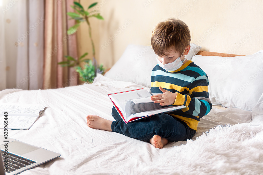 Distance learning online education. Quarantine boy with book and laptop in medical mask studying at home and doing school homework. Sitting on bed with training books. Distance learning online education. Quarantine boy with book and laptop in medical mask studying at home and doing school homework. Sitting on bed with training books.