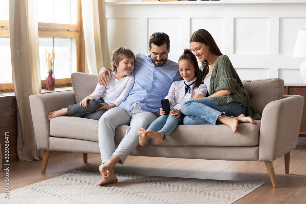Full length happy young family couple relaxing on comfortable sofa with smiling two little children siblings, using smartphone. Joyful parents spending free weekend time with kids in living room. Full length happy young family couple relaxing on comfortable sofa with smiling two little children siblings, using smartphone. Joyful parents spending free weekend time with kids in living room.