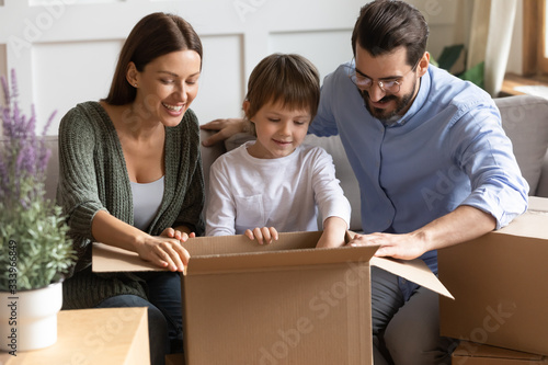 Happy little kid boy sitting on sofa between smiling parents, unboxing family belongings together after moving in new apartment. Joyful family of three unpacking cardboard boxes with stuff at home.