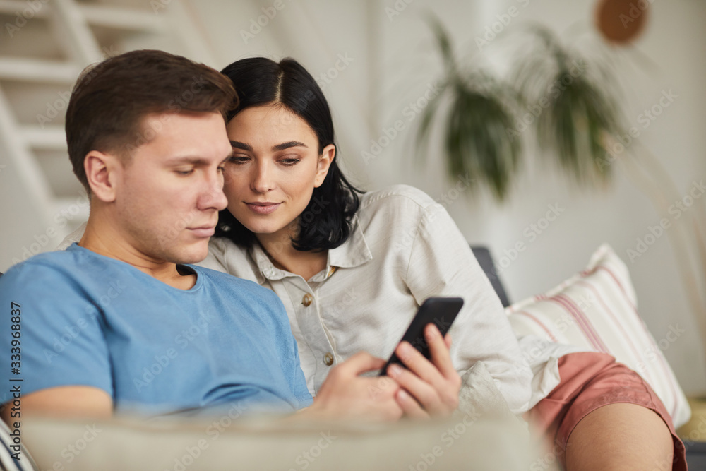 Fototapeta premium Portrait of modern young couple using smartphone while relaxing on sofa in cozy home interior, copy space