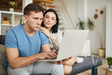 Portrait of modern young couple using laptop together while sitting on sofa in cozy home interior, online service concept, copy space