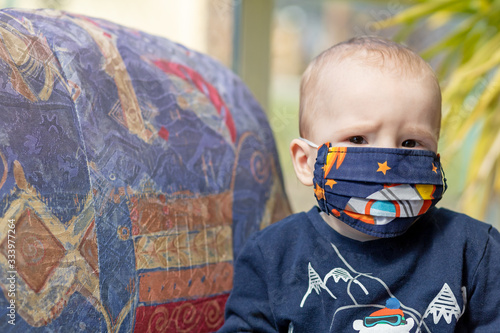 Portrait of angry baby with reusable protective face mask against coronavirus on face. The boy is looking at the camera. 