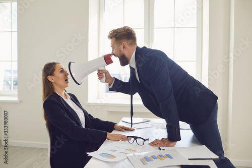 Boss businessman shouting in magaphone at businesswoman employee secretary at desk in office photo