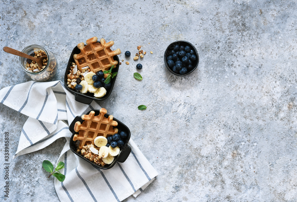 Belgian waffles with yogurt, granola, banana and blueberry for breakfast on a concrete background. View from above. Belgian waffles with yogurt, granola, banana and blueberry for breakfast on a concrete background. View from above.