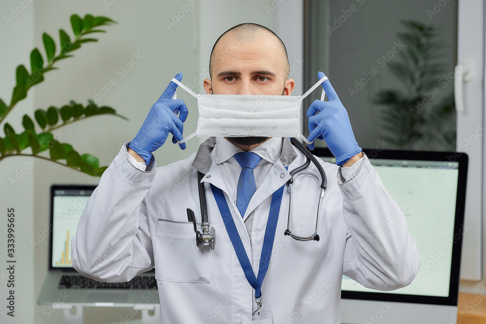 A caucasian doctor putting on a protective face mask to avoid the spread coronavirus (COVID-19) in his office. A bald physician with a beard preparing to examine a patient. A caucasian doctor putting on a protective face mask to avoid the spread coronavirus (COVID-19) in his office. A bald physician with a beard preparing to examine a patient.