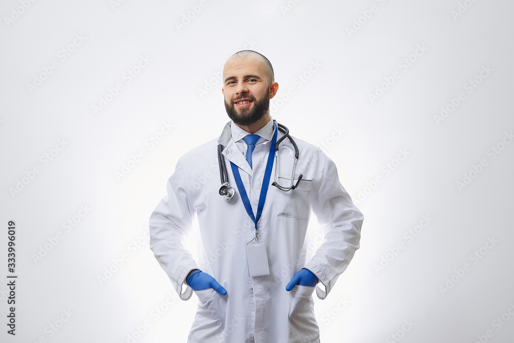 A happy doctor in disposable medical gloves with a stethoscope holding hands in pockets. A bald physician with a beard preparing to examine a patient.