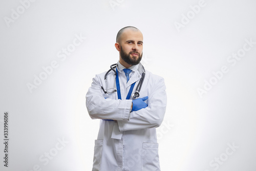 A doctor with arms crossed on his chest in disposable medical gloves with a stethoscope. A bald physician with a beard preparing to examine a patient.