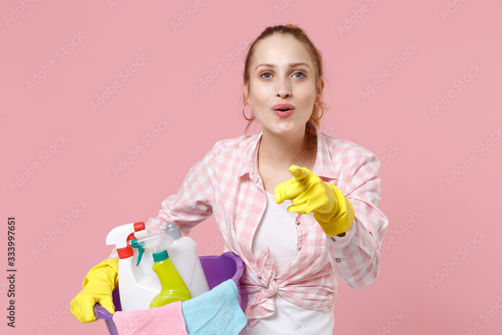 Cheerful young woman housewife in rubber gloves hold basin with detergent bottles washing cleansers doing housework isolated on pink background. Housekeeping concept. Pointing index finger on camera. Cheerful young woman housewife in rubber gloves hold basin with detergent bottles washing cleansers doing housework isolated on pink background. Housekeeping concept. Pointing index finger on camera.