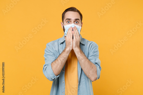 Shocked young man in sterile face mask posing isolated on yellow background studio portrait. Epidemic pandemic spreading coronavirus 2019-ncov sars covid-19 flu virus concept. Cover mouth with hands.