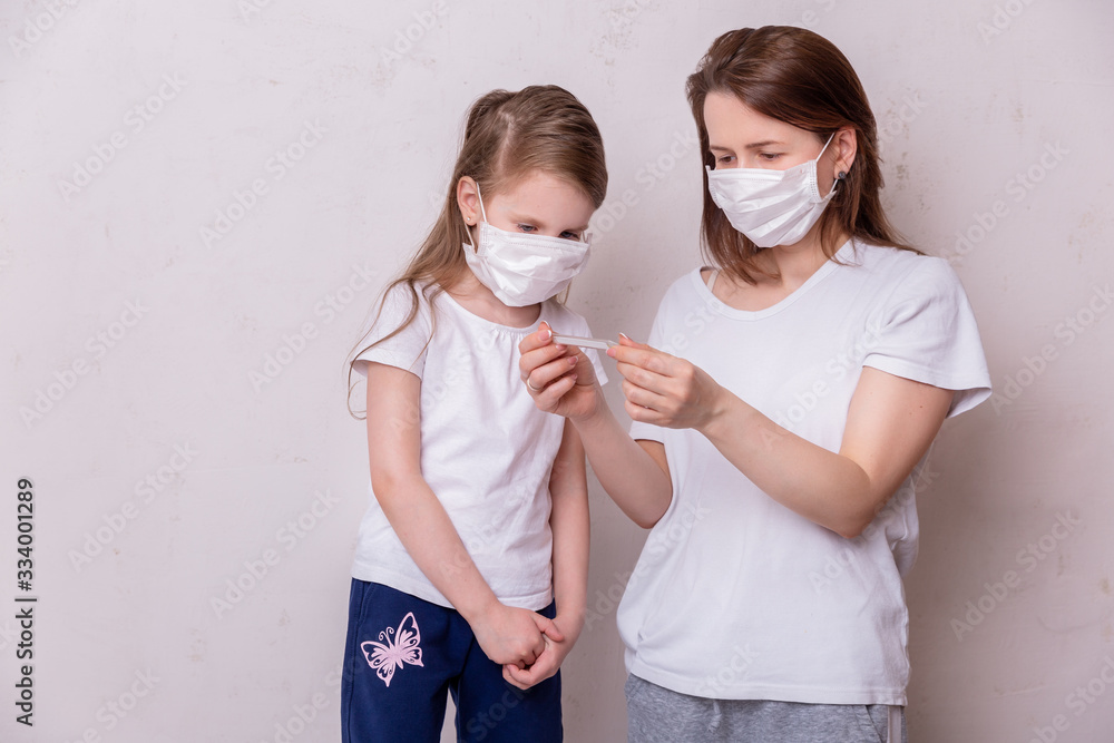 The concept of coronavirus quarantine. Young woman and her daughter in medical masks in isolation at home. Mom measures the temperature of her daughter. Theme of health and medicine