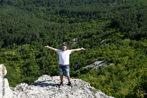 A handsome young man stands on the edge of a cliff and spreads his arms in different directions, behind a beautiful forest panorama. The man is happy and pleased with the beautiful beauties of nature.