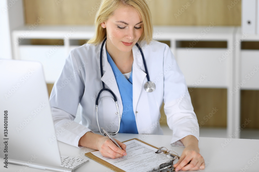 Doctor woman at work while sitting at the desk in hospital or clinic. Blonde cheerful physician filling up medical records form. Data and best service in medicine and healthcare Doctor woman at work while sitting at the desk in hospital or clinic. Blonde cheerful physician filling up medical records form. Data and best service in medicine and healthcare