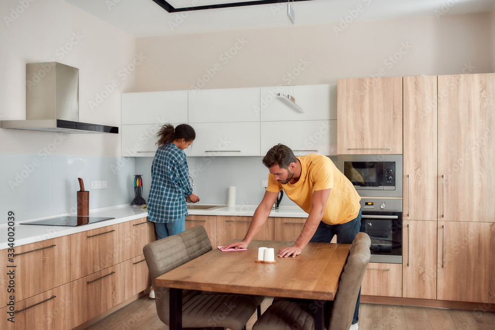 Let's work together. Young couple in casual clothes cleaning kitchen together. Caucasian man cleaning kitchen table, while afro american woman washing dishes on the background Let's work together. Young couple in casual clothes cleaning kitchen together. Caucasian man cleaning kitchen table, while afro american woman washing dishes on the background