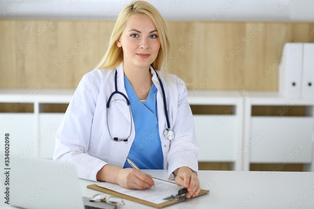Doctor woman at work while sitting at the desk in hospital or clinic. Blonde cheerful physician filling up medical records form. Data and best service in medicine and healthcare Doctor woman at work while sitting at the desk in hospital or clinic. Blonde cheerful physician filling up medical records form. Data and best service in medicine and healthcare