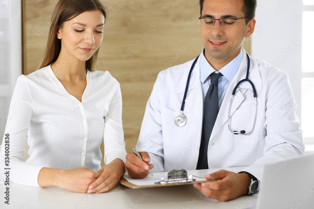 Doctor and patient discussing the results of a physical examination while sitting at a desk in a clinic. A male doctor using a clipboard to fill out a medical history of a young woman's medication Doctor and patient discussing the results of a physical examination while sitting at a desk in a clinic. A male doctor using a clipboard to fill out a medical history of a young woman's medication