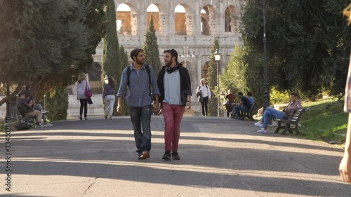 Young happy gay couple tourists walk in park road with trees colosseum in background in rome at sunset holding hands lovely slow motion colle oppio