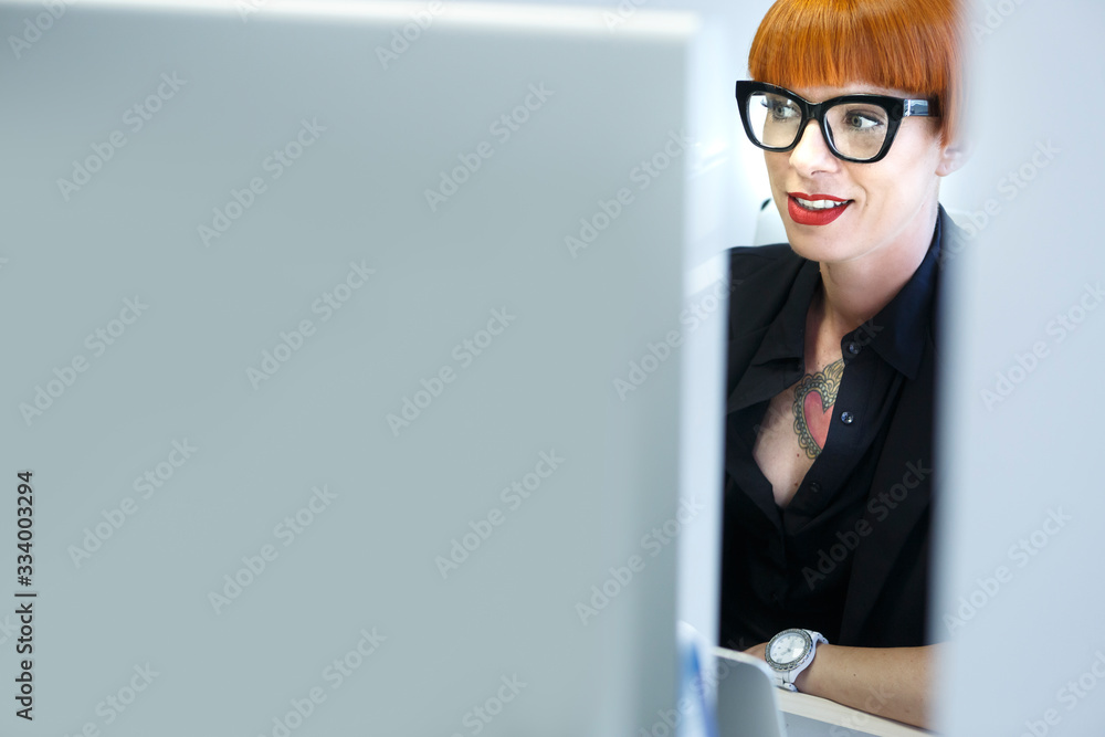 Manager con i capelli rossi a caschetto, tatuaggi, giacca nera e gli occhiali neri sta nella sua postazione di fronte a un monitor Manager con i capelli rossi a caschetto, tatuaggi, giacca nera e gli occhiali neri sta nella sua postazione di fronte a un monitor