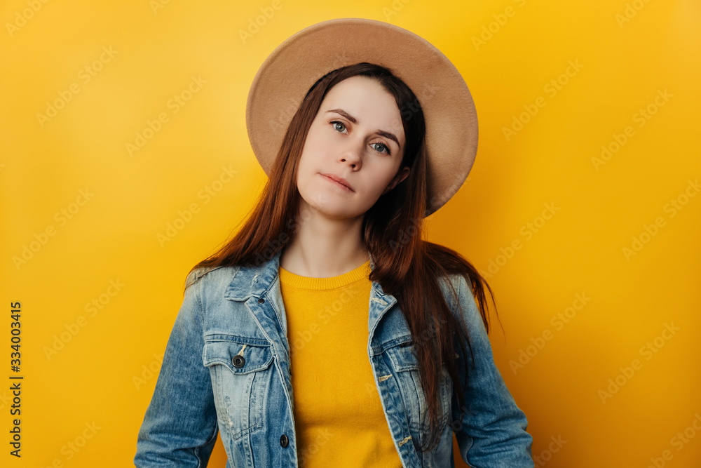 Attractive young woman in hat, calm smile, dressed in casual blue denim jacket, looks straight at camera, pose over yellow studio background with copy space. Human emotions and feelings concept Attractive young woman in hat, calm smile, dressed in casual blue denim jacket, looks straight at camera, pose over yellow studio background with copy space. Human emotions and feelings concept