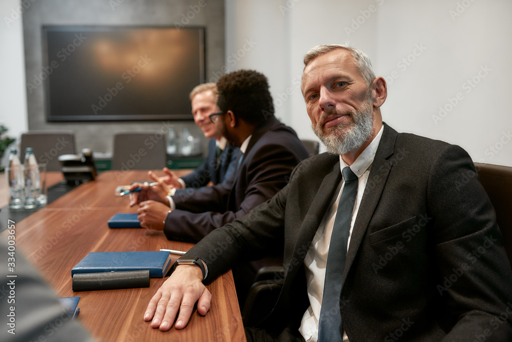 Portrait of bearded mature man in formal wear looking at camera while sitting at the office table with his coworkers in the modern office Portrait of bearded mature man in formal wear looking at camera while sitting at the office table with his coworkers in the modern office