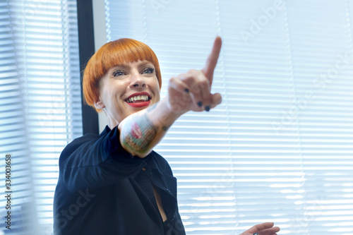 Manager donna con i capelli rossi a caschetto e i tatuaggi, vicino a una veneziana punta il dito ridendo