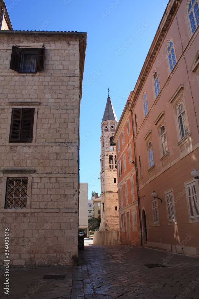 Narrow streets of a European city with red roofs and stone walls. A platform of stone and a shadow in the city. A chapel in the distance among tall houses.