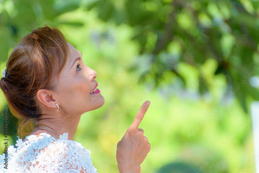 Side face of a beautiful woman healthy middle-aged Asian people dress in Thai style clothes, Portrait old lady relaxing outdoors pointing finger looking up in the park, copy space on nature background Side face of a beautiful woman healthy middle-aged Asian people dress in Thai style clothes, Portrait old lady relaxing outdoors pointing finger looking up in the park, copy space on nature background