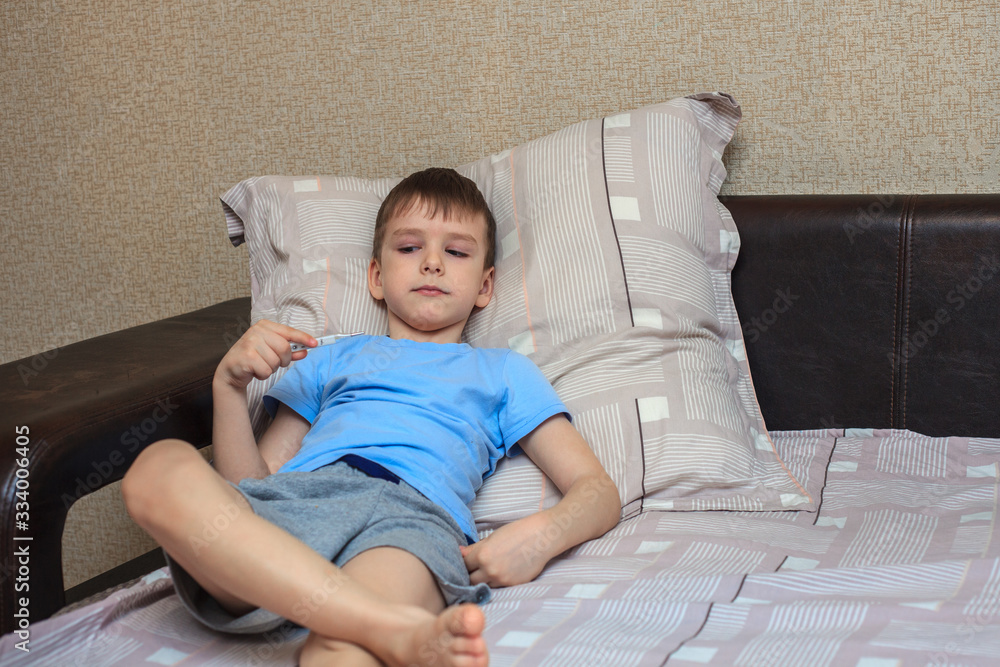 The child lies on the bed with a thermometer in his hands, measuring temperature. Virus, flu. Home quarantine The child lies on the bed with a thermometer in his hands, measuring temperature. Virus, flu. Home quarantine