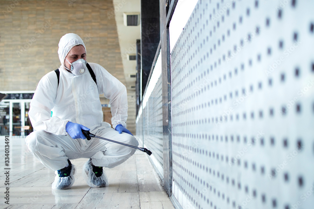 Wall Mural Shot of male person in white chemical protection suit doing disinfection of public areas to stop spreading highly contagious corona virus