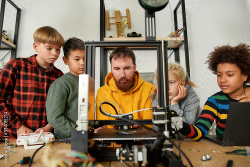 Experience digital innovation. Group of smart kids listening to their male teacher while looking at 3d printer and plastic detail prototype at robotics school lesson photo