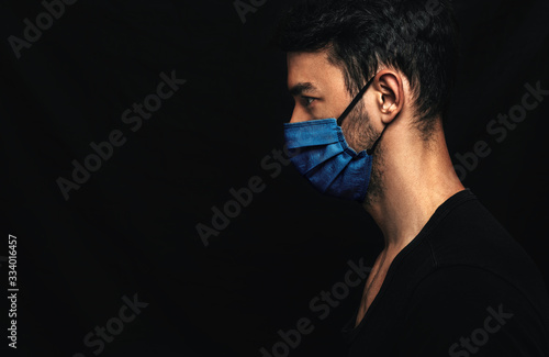 Side view image of Caucasianman wearing medical blue mask on the face during virus pandemic lockdown posing on the black wall with copy space. Male with disposable face mask to prevent the coronavirus photo