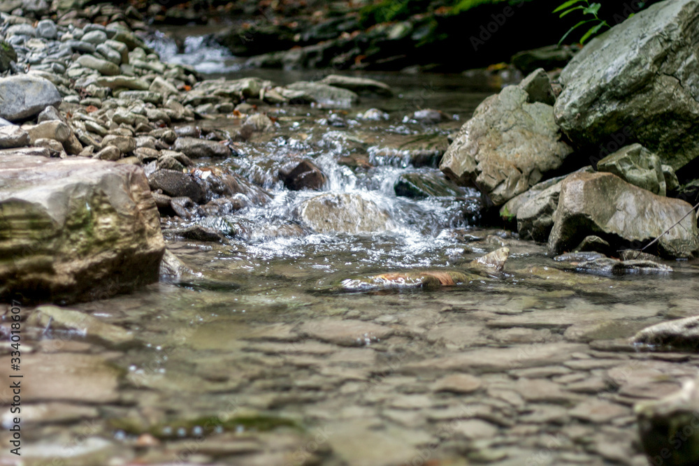 A small waterfall in a mountain stream in the Caucasus Mountains. View of the waterfall on a summer day. Algae grow on stons within a stream bed. Stones under water. A small waterfall in a mountain stream in the Caucasus Mountains. View of the waterfall on a summer day. Algae grow on stons within a stream bed. Stones under water.