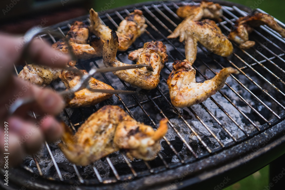 Grilling at summer weekend. Fresh meat preparing on grill. Lifting chicken wings with pair of tongs. Assorted delicious grilled meat.