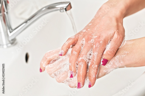 Young woman washing her hands under water tap faucet with soap. Detail on fingers, nails covered purple polish. Personal hygiene concept - coronavirus covid-19 outbreak prevention