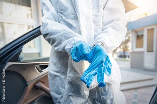 Doctor, scientist or nurse wearing face mask and biological hazmat protective suit putting on rubber gloves near car outdoor. Coronavirus covid-19 outbreak spread alert danger