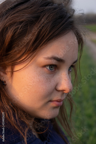 Girl face with freckles on the side against the background of green and bright evening sunlight.