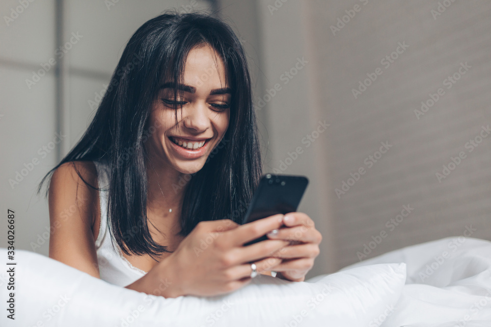 Happy Beautiful woman texting at smartphone while lying on her bed. Smiling Brunette Girl holding mobile phone Happy Beautiful woman texting at smartphone while lying on her bed. Smiling Brunette Girl holding mobile phone