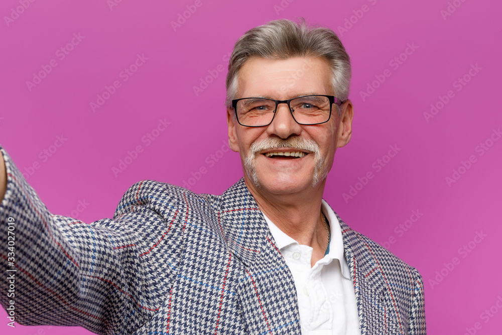 Studio portrait of happy senior male in eyeglasses smiling while posing in violet background, selfie time Studio portrait of happy senior male in eyeglasses smiling while posing in violet background, selfie time