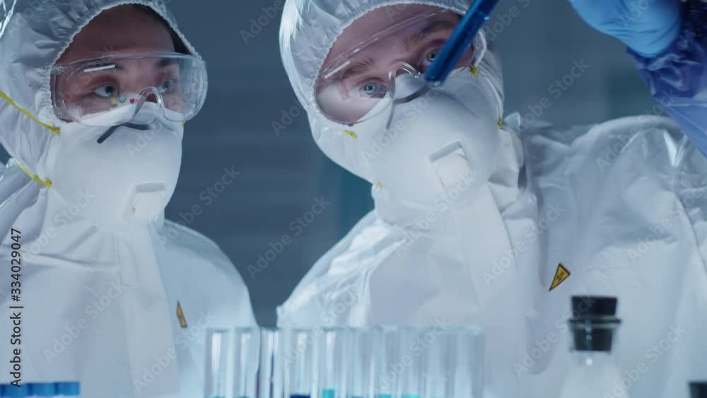 Tilt up shot of male and female multiethnic scientists s in protective apparel examining blue liquid in test tube and talking while working together on antiviral vaccine in laboratory Tilt up shot of male and female multiethnic scientists s in protective apparel examining blue liquid in test tube and talking while working together on antiviral vaccine in laboratory