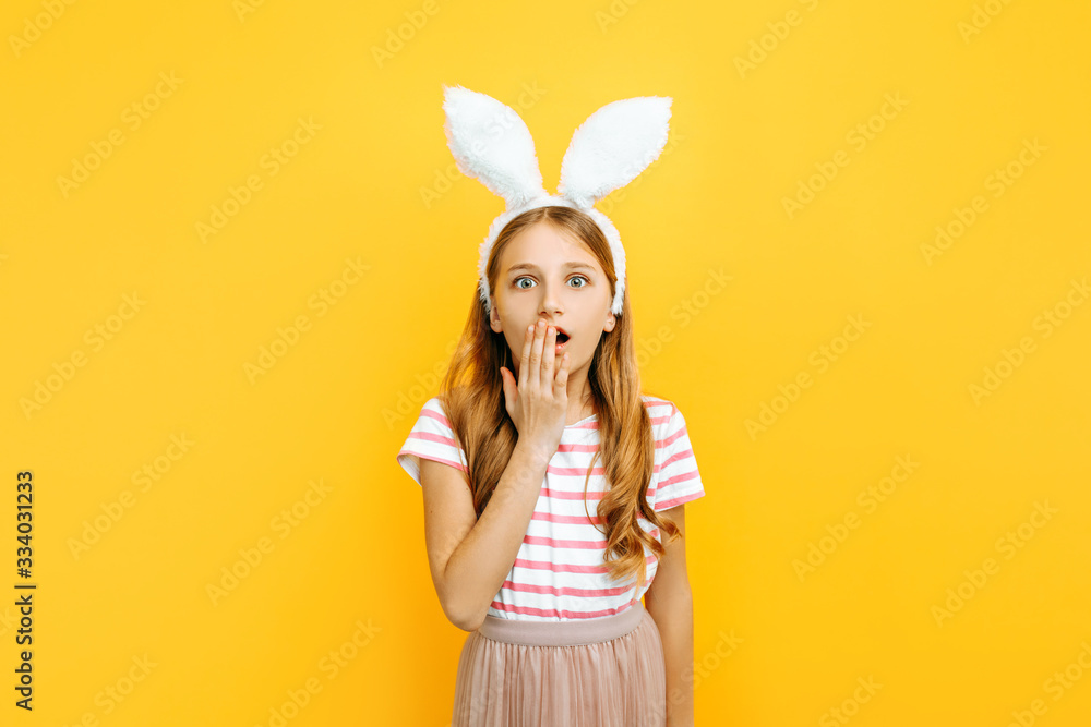 Shocked little beautiful girl on the head with rabbit ears, surprised covers her mouth with her hand on a yellow background